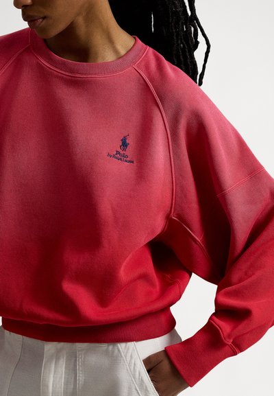 Rød genser med gradientdesign, med ribbet rund hals, raglanermer og brodert logo i blått. Myk stofftekstur.