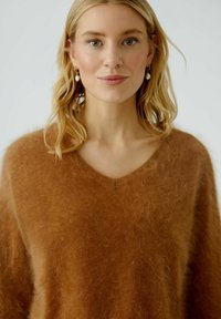 Brauner, flauschiger Pullover mit V-Ausschnitt. Weicher, strukturierter Stoff. Das Model hat lockige Haare und trägt goldene Ohrringe.