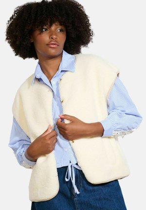 Gilet in pile color crema con orlo arrotondato e bottoni dorati, indossato sopra una camicia a righe blu e bianche con dettagli in pizzo.