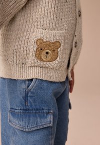 Cardigan beige en tricot avec un patch ours sur la poche gauche, associé à un pantalon cargo en denim bleu avec poches latérales et détails de coutures.