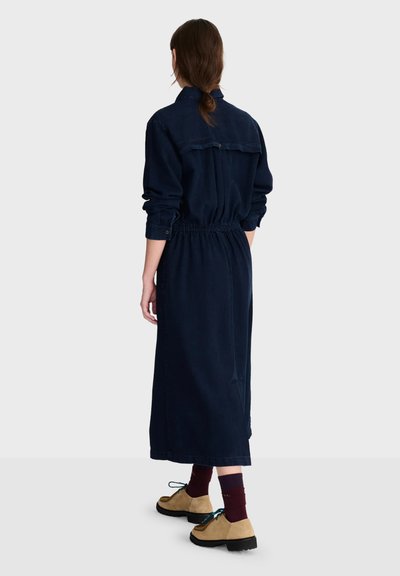 Femme aux cheveux bruns attachés porte une longue robe bleu marine avec une taille cintrée, des manches retroussées, des chaussettes bordeaux et des chaussures à lacets couleur beige.