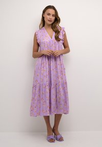 Cvečni vijolični oblek s V-izrezom, nabranimi rokavi in večplastnim krilom. Gladek material z oranžnimi in rumenimi cvetličnimi vzorci. Vijolične sandale.