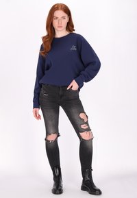 Felpa navy con vestibilità ampia e maniche lunghe, abbinata a jeans skinny neri strappati. Modello che indossa stivali neri alla caviglia.
