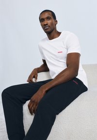 Biele bavlnené tričko s červeným logom "HUGO", v kombinácii s čiernymi textúrovanými nohavicami. Model sediaci na svetlom povrchu. Jednoduchý dizajn.