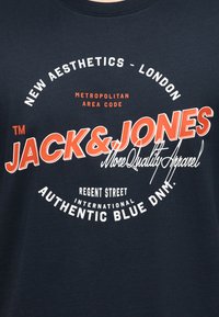 Ύφασμα σε ναυτικό μπλε με λευκό και πορτοκαλί κείμενο: "New Aesthetics - London, Metropolitan Area Code, Jack & Jones, More Quality Apparel, Regent Street, Authentic Blue DNM."
