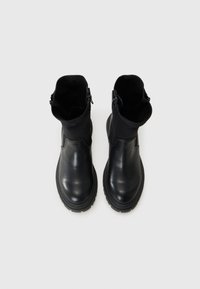 Bottines noires avec tiges en cuir lisse et en tissu, dotées de fermetures éclair latérales, posées sur un fond blanc uni.