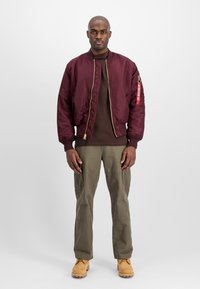Mies seisoo kasvot eteenpäin burgundin värisessä bomber-takissaan, ruskeassa paidassa, oliivinvihreissä cargo-housuissa ja vaaleissa työsaappaissa vaaleaa taustaa vasten.