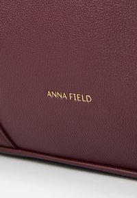 Burgundy handbag with textured surface, featuring gold "ANNA FIELD" logo. Includes sleek lines and a structured design.