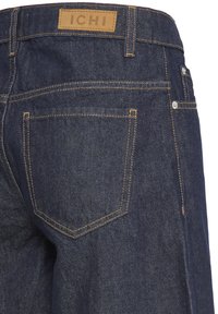 Mørkeblå denimjeans med baglomme, gule syninger og et tanfarvet lædermærke med mærkenavnet "ICHI" på linningen.