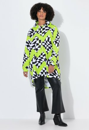 Femme portant un long sweat à capuche à motif zigzag noir, vert néon et blanc, avec un pantalon en simili cuir noir et des bottes à talons noires, se tenant devant un fond blanc.
