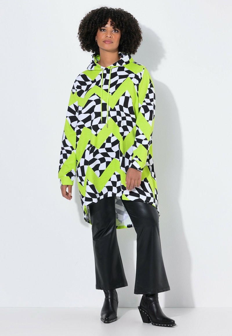 Donna che indossa un lungo hoodie a zigzag neon verde, nero e bianco, con pantaloni neri in faux pelle e stivali col tacco neri, in piedi contro uno sfondo bianco.