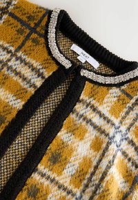 Strickjacke in Senfgelb, Schwarz und Grau im Karodesign mit texturierter Oberfläche; verfügt über einen schwarzen Kragen, der mit silbernen Perlen und einem Etikett verziert ist.