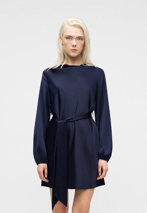 Ξανθιά γυναίκα με μακρυμάνικο φόρεμα σε σκούρο μπλε χρώμα, με ζώνη, στέκεται μπροστά σε απλό ανοιχτό γκρι φόντο.