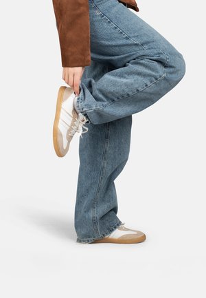 Persona che indossa jeans blu e giacca marrone solleva una gamba, tenendo la caviglia per regolare delle sneakers bianche e beige con suola in gomma.