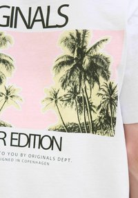 Hvid bomulds t-shirt med et palmetræ grafik i grønne og gule nuancer mod en lyserød baggrund, med fed sort tekst ovenfor og nedenfor.