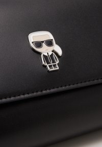 Musta nahkalompakko, jossa on sarjakuvapinssi aurinkolaseja käyttävästä hahmosta, jolla on valkoiset hiukset ja musta puku. Saumaus yksityiskohtia reunan ympärillä.