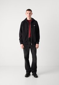 Černá mikina na zip s šňůrkami přes vínové triko, v kombinaci s tmavými džíny a černými botami, které nosí stojící model.
