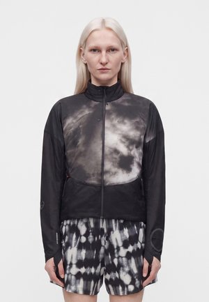 Ženska z dolgimi blond lasmi nosi črno-belo jopico z vzorcem oblakov z zadrgo in črno-bele kratek hlače z vzorcem tie-dye na enobarvnem ozadju.