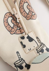 Sac en toile avec un fond crème présentant des motifs de bretzels et d'imprimés vache. Il est doté d'une fermeture éclair beige et d'une languette pour la fermeture.