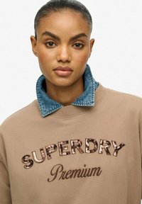 Beżowa bluza z napisem "SUPERDRY Premium" wyszytym cekinami. Posiada kontrastowy kołnierz z denimu. Gładka faktura, swobodny krój, luźny fason.