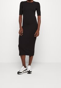 Robe noire midi à manches courtes, design ajusté, texture lisse, associée à des baskets athlétiques blanches et noires.