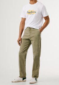 Homme portant un t-shirt blanc Pepe Jeans London, un pantalon vert olive et des baskets blanches avec des détails verts, debout avec une main dans la poche.