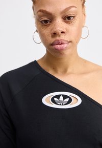 Svart ribbet topp med avskåret skulderdesign og oval utskjæring med hvit brodert logo. Glatt tekstur med ringøredobber.