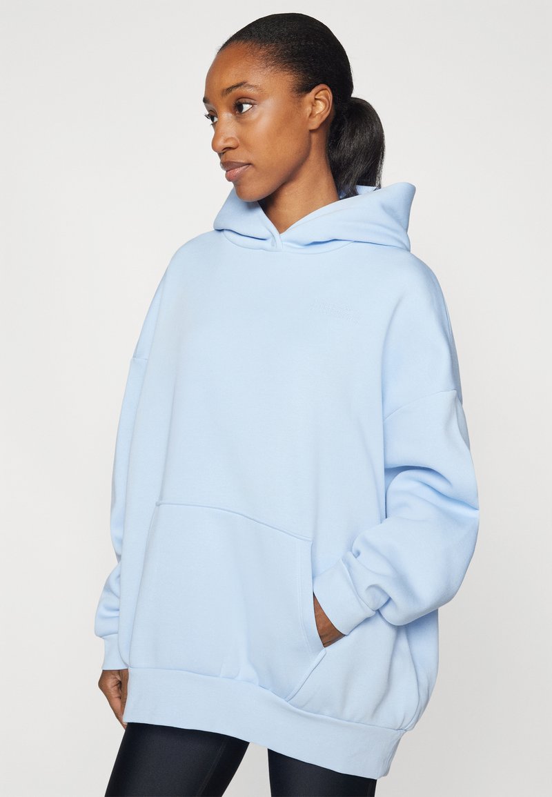 Frau mit dunklem Pferdeschwanz, die einen übergroßen, hellblauen Hoodie mit Taschen trägt und seitlich vor einem schlichten Hintergrund schaut.