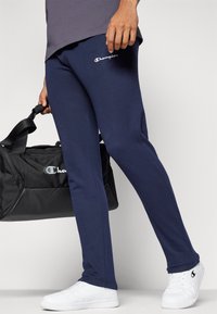 Marineblaue Sweatpants aus weichem Stoff, mit seitlichen Taschen und einem kleinen Champion-Logo auf dem Oberschenkel. Kombiniert mit einer schwarzen Sporttasche.