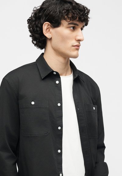 Νέος με σγουρά σκούρα μαλλιά, φορώντας ένα μαύρο πουκάμισο με κουμπιά πάνω από ένα λευκό T-shirt, κοιτάζοντας προς τα δεξιά μπροστά σε απλό φόντο.