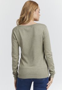 Olivenfarget strikkegenser med lange ermer, bred utringning og ribbede mansjetter og kant, kombinert med mørkeblå jeans.