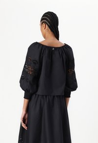 Top nero senza spalle con maniche larghe e texture che presentano dettagli ricamati. Tessuto morbido con vita arricciata e chiusura con bottone sul retro.