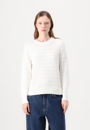 Hvit strikket genser med horisontale striper, rund halslinning og knappedetalj på skulderen. Kombinert med blå denimbukser.