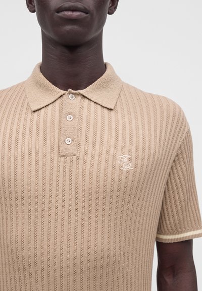 Haut du corps d’une personne portant un polo beige en maille côtelée avec un col texturé et trois boutons, affichant un petit logo brodé.