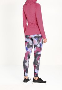 Maglia sportiva rossa a maniche lunghe con cappuccio, abbinata a leggins multicolori a fantasia in nero, rosa, viola e grigio. Scarpe da ginnastica nere.