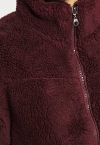 Veste en polaire bordeaux avec col montant, fermeture éclair intégrale à l'avant, et une surface douce et texturée. Comprend un détail de couture horizontale.