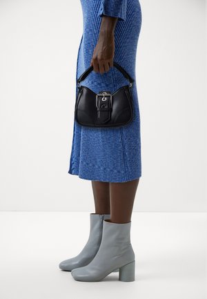 Una persona che indossa un vestito blu a coste tiene una piccola borsa in pelle nera e indossa stivaletti grigio chiaro con tacchi a blocco.