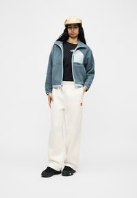 Γυναίκα που φοράει μπεζ καπέλο, μπλε φλις μπουφάν με φερμουάρ, μαύρο μπλουζάκι, άσπρο φαρδύ παντελόνι και μαύρα παπούτσια, στέκεται με το ένα χέρι στην τσέπη.