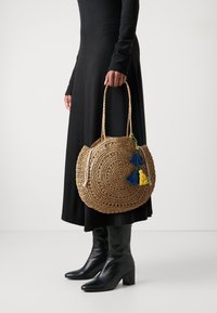 Sac rond en paille avec une texture tissée, ornée de pompons bleus et jaunes. Le sac dispose de deux poignées solides et est de couleur naturelle.