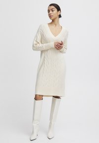 Hellbeige Strickkleid mit Kabelmuster, V-Ausschnitt, langen Ärmeln und geripptem Saum. Kombiniert mit knielangen weißen Absatzstiefeln.
