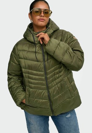Žena so zelenou oversized nadýchanou bundou s kapucňou, modrými džínsami a veľkými tónovanými slnečnými okuliarmi, ktorá drží golier bundy jednou rukou.