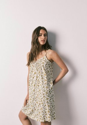 Donna con lunghi capelli castani che indossa un vestito bianco senza maniche con motivo floreale giallo, in piedi contro un muro chiaro e uniforme.