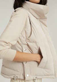 Jasnobeżowa pikowana kamizelka z wysokim kołnierzem, boczną kieszenią, zatrzaskami i regulowanym dołem. Pikowany wzór o gładkim wykończeniu.