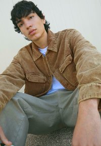 Giacca in corduroy color marrone chiaro con due tasche frontali, abbinata a una camicia bianca e pantaloni grigi, seduto su una superficie testurizzata.