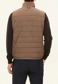 Braune gesteppte Weste mit hohem Kragen, horizontaler Steppung und Seitenknöpfen, getragen über einem dunklen Langarmshirt und navyfarbenen Hosen.