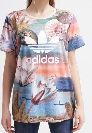 Person iført en farverig Adidas t-shirt med store blomsterprint og en solnedgangsbaggrund, med det hvide Adidas-logo midt på.