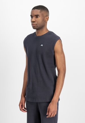 Junger Mann steht und trägt ein ärmelloses, dunkelgraues, geripptes Shirt mit kleinem Logo auf der Brust vor einem schlichten hellen Hintergrund.