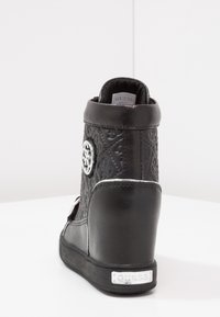 Musta viistokärkinen sneaker, jossa on teksturoitu nahka, hopeiset yksityiskohdat ja koristeellinen embleemi. Varustettu pehmeällä kauluksella ja kestävällä kumipohjalla.