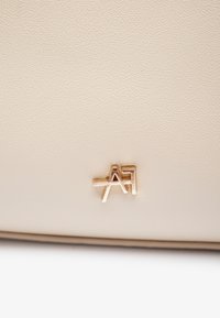 Beige teksturert håndveske med en gylden metalllogo festet på forsiden. Enkel design med rene linjer og minimalistiske detaljer.