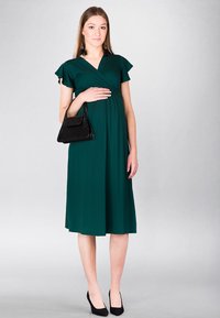 Ciemnozielona sukienka ciążowa do kolan z krótkimi falbanastymi rękawami, z dekoltem w serek, noszona z czarną torebką i czarnymi szpilkami.